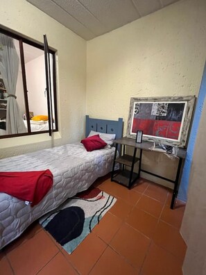 Ein kleines, gemütliches Zimmer mit einem Einzelbett, einem Schreibtisch mit Computer und einem Fenster mit Vorhängen.