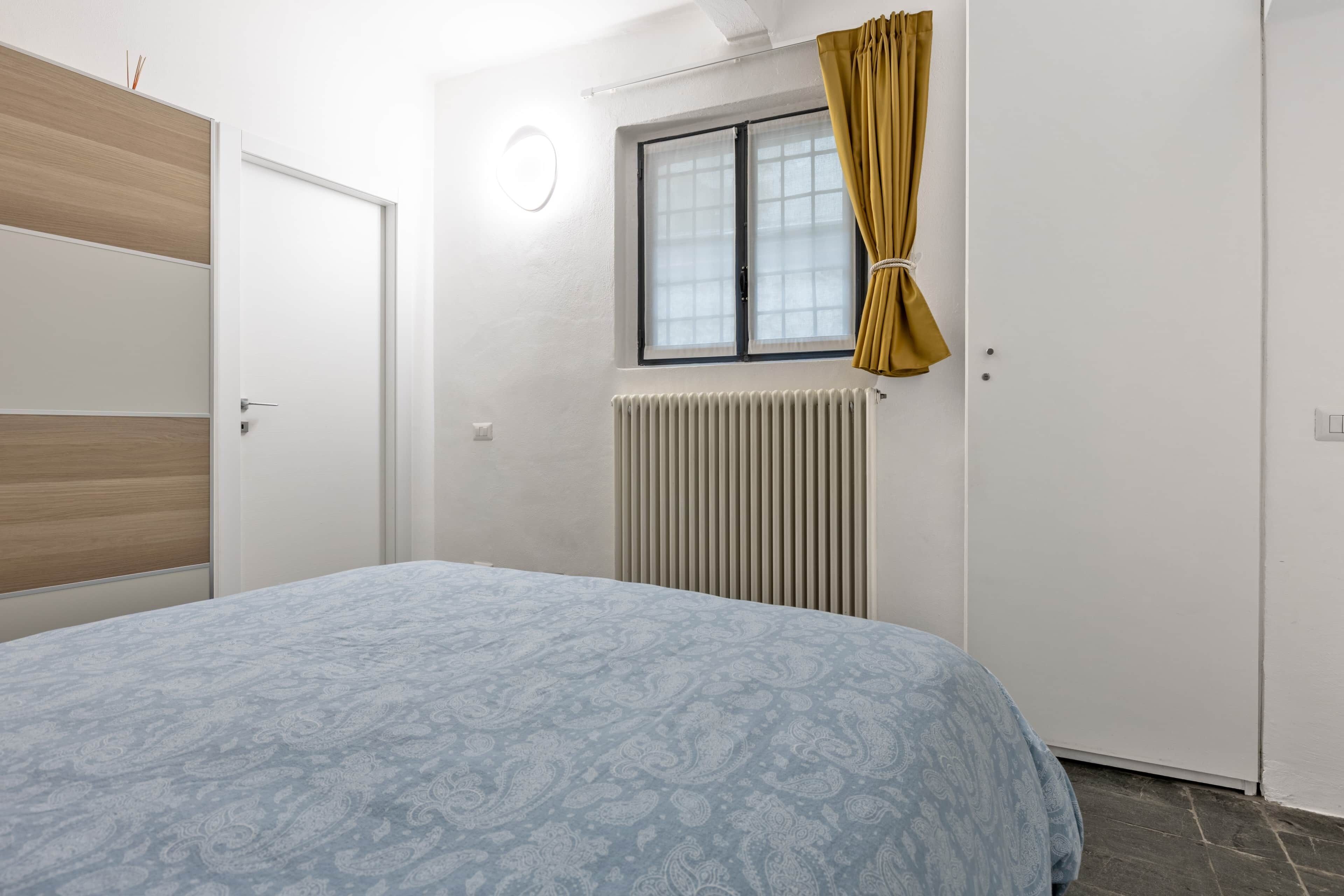 A bedroom with a bed, a window with curtains, a radiator, and a door.