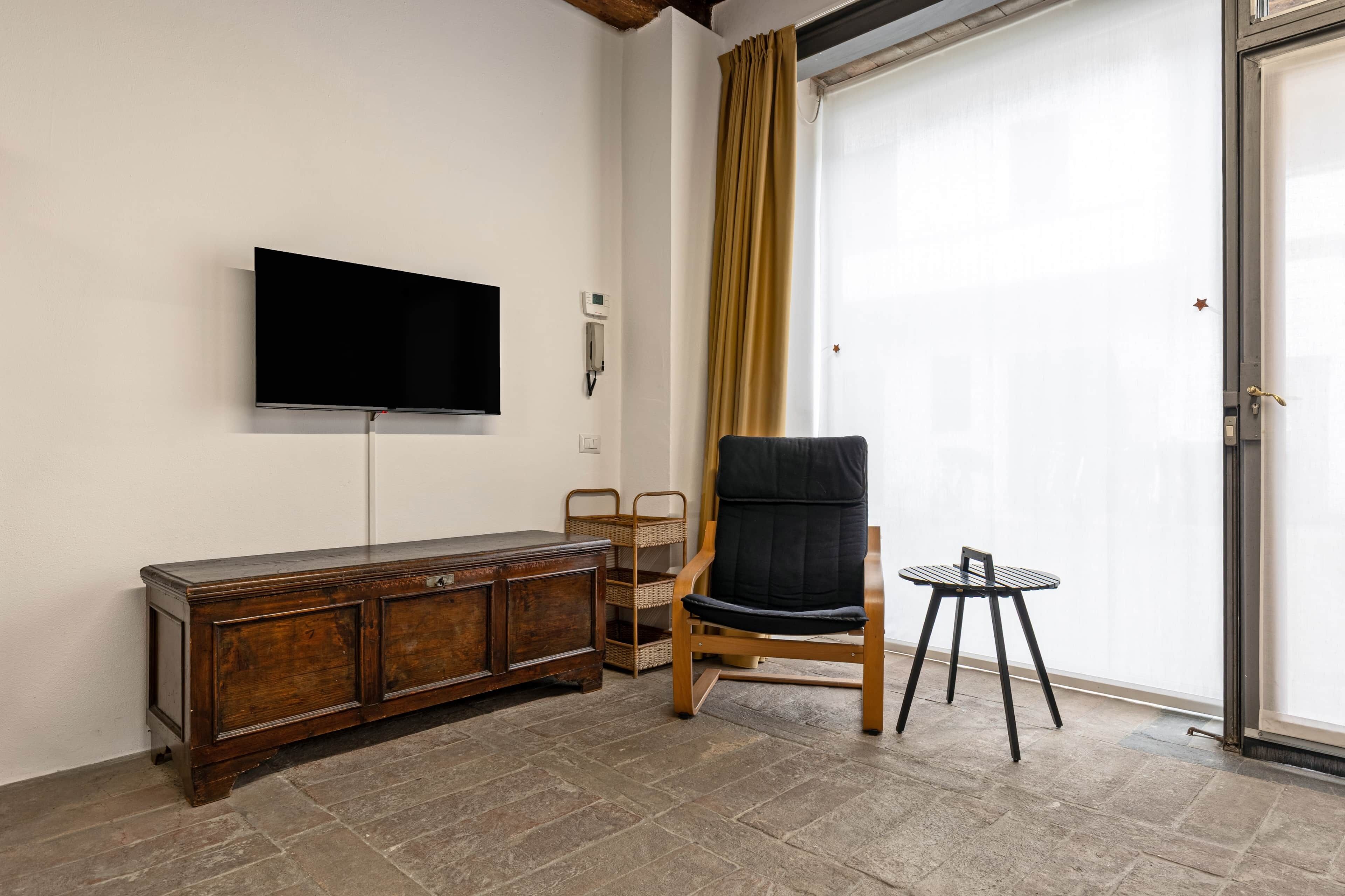 A room with a wooden cabinet, a black chair, a small round table, a television mounted on the wall, and a sliding glass door with a curtain.