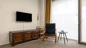 A room with a wooden cabinet, a black chair, a small round table, a television mounted on the wall, and a sliding glass door with a curtain.