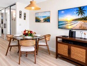 A dining area with a round table, four chairs, a vase of flowers, and a television on a wooden cabinet. - Papakea | 3 Ocean View Villas, Sleeps 12 | Car Incl. w/ 6+ Nights | ML-4262 by KBM (Lahaina)