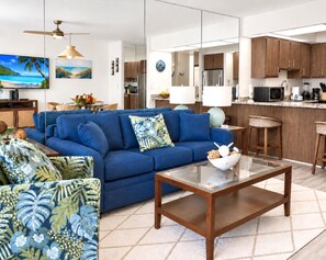 Ein modernes Wohnzimmer mit einem blauen Sofa, einem Glastisch und einer Küchenzeile mit Holzschränken und einer Mikrowelle.