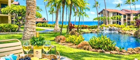 A tropical resort with a pool, palm trees, and a dining area with a basket of fruit and wine glasses.