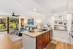 A modern kitchen with a central island, a dining area with a blue sofa, and a view of a landscaped garden. - Papakea | 3 Ocean View Villas, Sleeps 12 | Car Incl. w/ 6+ Nights | ML-4262 by KBM (Lahaina)