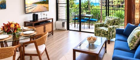 A modern living room with a dining area, a large TV, and a view of a tropical landscape through the balcony doors.
