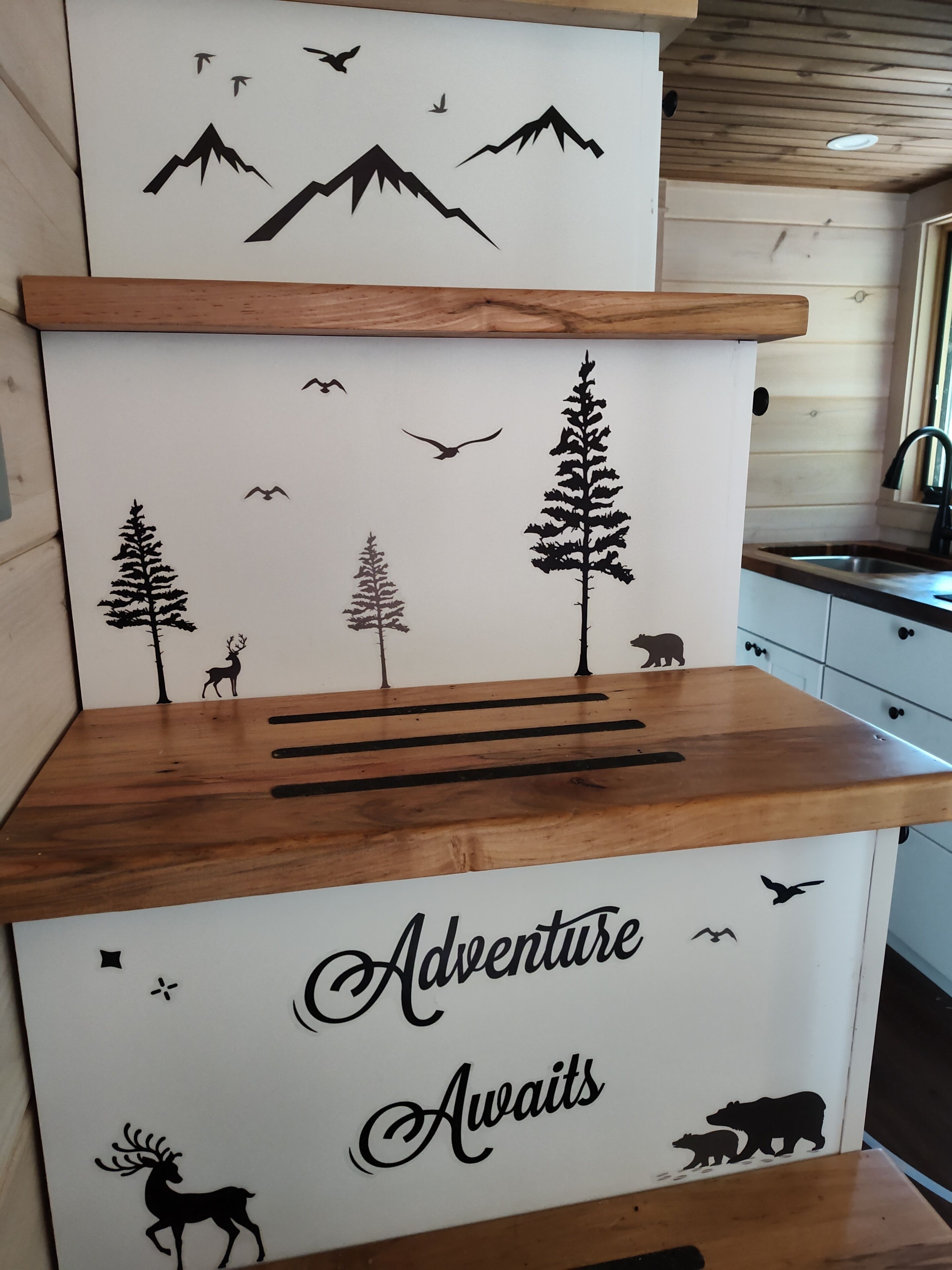 Una cocina con gabinetes de madera, una encimera y un adhesivo de pared con una escena natural que muestra montañas, árboles y siluetas de animales.
