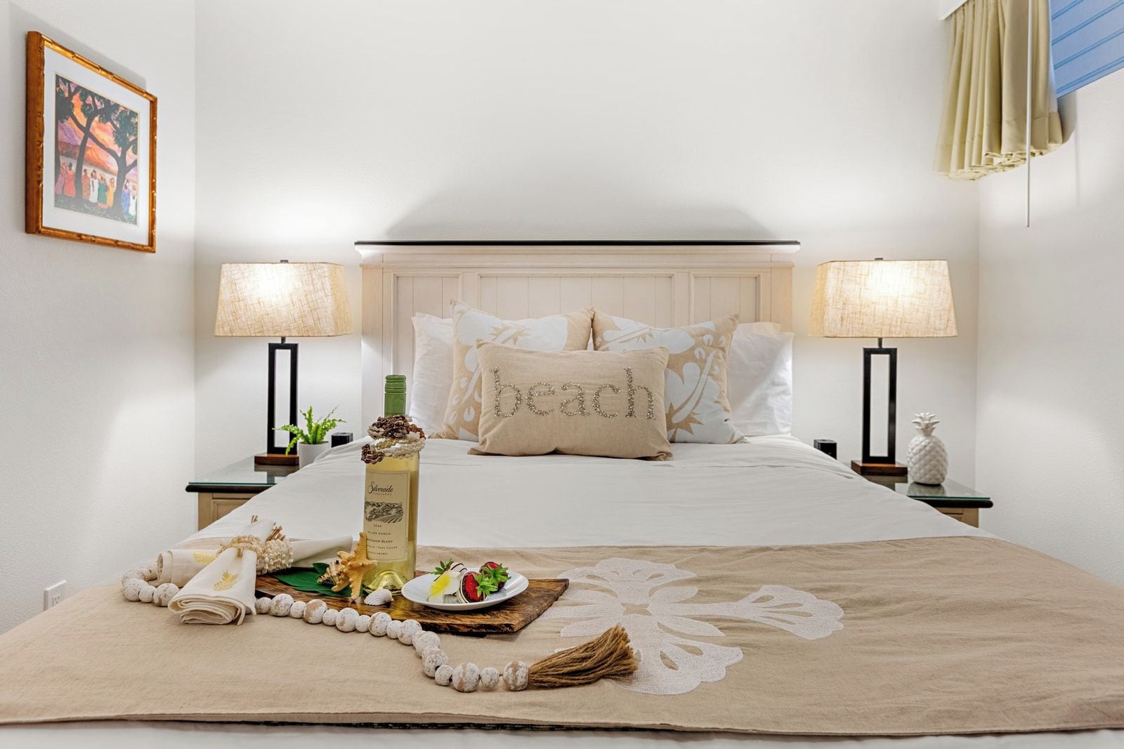 A neatly arranged bedroom with a bed, two bedside lamps, a bedside table with a decorative item, a framed picture on the wall, and a decorative pillow with the word 'beach' on it.