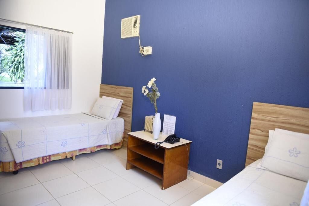 Una habitación con dos camas, una mesita de noche con teléfono y un florero, una ventana con cortinas y un aire acondicionado instalado en la pared.
