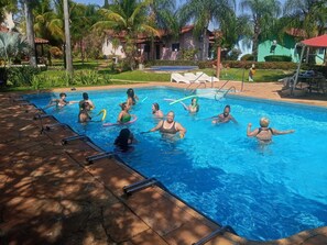 A swimming pool with people in it, surrounded by palm trees and a building with a red roof. - Rosa Spa e Pousada (Aparecida de Goiânia)