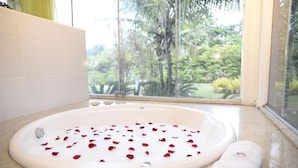 A bathroom with a bathtub filled with rose petals, a towel, and a view of a tropical garden through a window.