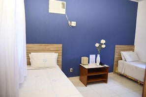 A bedroom with a blue wall, a wooden headboard, a white bed with a floral pattern, a bedside table with a vase and a lamp, and a small wooden nightstand. - Rosa Spa e Pousada (Aparecida de Goiânia)