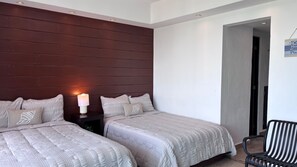 Habitación de hotel con dos camas, una mesita de noche con lámpara, una silla y una puerta que da a otra habitación.