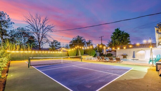 Un court de tennis extérieur avec un filet, entouré de guirlandes lumineuses, et un espace piscine avec des chaises longues.