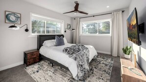 Une chambre à coucher avec un lit, un ventilateur de plafond, deux fenêtres, une table de chevet, un téléviseur et un tableau.