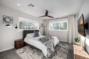 A bedroom with a bed, a ceiling fan, two windows, a nightstand, a TV, and a painting. - Pickleball Ball Paradise (Phoenix)