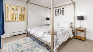 Una cama con dosel, con una colcha de diseño de mármol, dos mesitas de noche con lámparas y cuadros en las paredes.