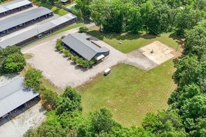 An aerial view of a property with a large covered parking area, a fenced sports field, and surrounding greenery. - The Pickwick Perch | Cozy 2BD Near Lake (Counce)