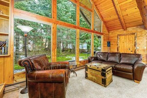 A log cabin interior with a leather sofa, a coffee table, and large windows offering a view of a forest.