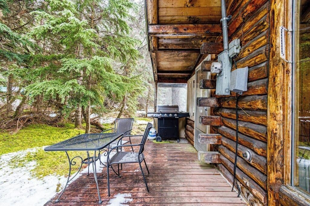 A wooden cabin with a deck, a grill, and a table with chairs.