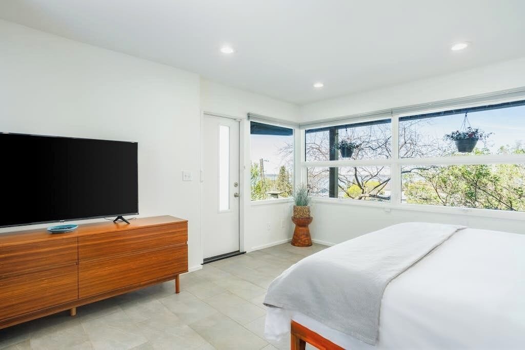 A bedroom with a bed, a television on a wooden dresser, and a window with a view of trees.