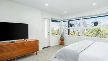 A bedroom with a bed, a television on a wooden dresser, and a window with a view of trees.