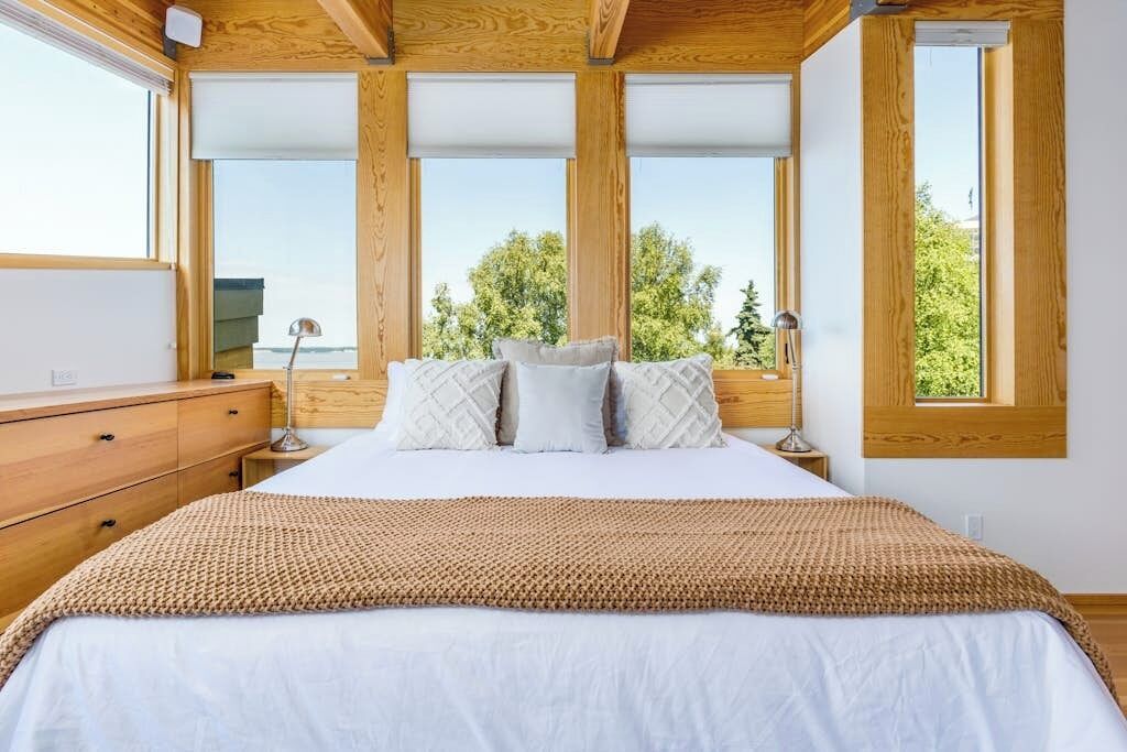 A bedroom with a large bed, wooden headboard, and a view of trees through large windows.