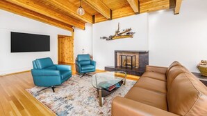 A living room with wooden ceiling beams, a fireplace, a mounted TV, and two blue armchairs. - Full Downtown Anchorage Triplex for Large Groups (Anchorage)