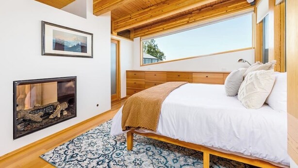 A bedroom with a wooden ceiling, a bed with white bedding, a fireplace, and a large window. - Full Downtown Anchorage Triplex for Large Groups (Anchorage)
