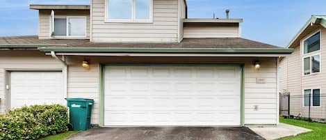 A two-story residential building with a garage door, a small attached garage, and a green trash bin.