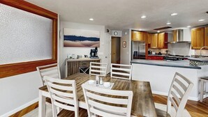 A dining area with a wooden table and white chairs, a kitchen with wooden cabinets and stainless steel appliances, and a painting on the wall. - Elegant Anchorage Lakehouse w/ Direct Water Access (Anchorage)