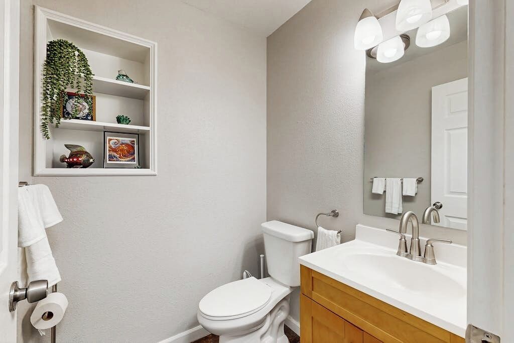 A bathroom with a toilet, sink, and mirror. There is a shelf with decorative items and a hanging plant.