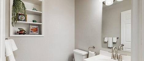 A bathroom with a toilet, sink, and mirror. There is a shelf with decorative items and a hanging plant.
