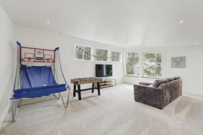 A living room with a foosball table, a sofa, a TV, and large windows. - Contemporary New-build Home With Lower Game Room (Girdwood)