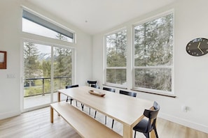 A dining area with a long table, chairs, and a bench. There are large windows with a view of trees and mountains. A clock is on the wall.