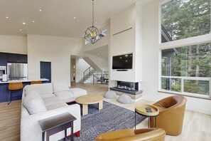 A spacious living area with a modern fireplace, a large TV, and a view of the outdoors. - Contemporary New-build Home With Lower Game Room (Girdwood)