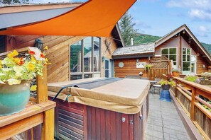 A wooden cabin with a hot tub, outdoor seating, and a large window overlooking a mountainous landscape. - Glacier-view Studio w/ Its Own Private Entrance (Girdwood)