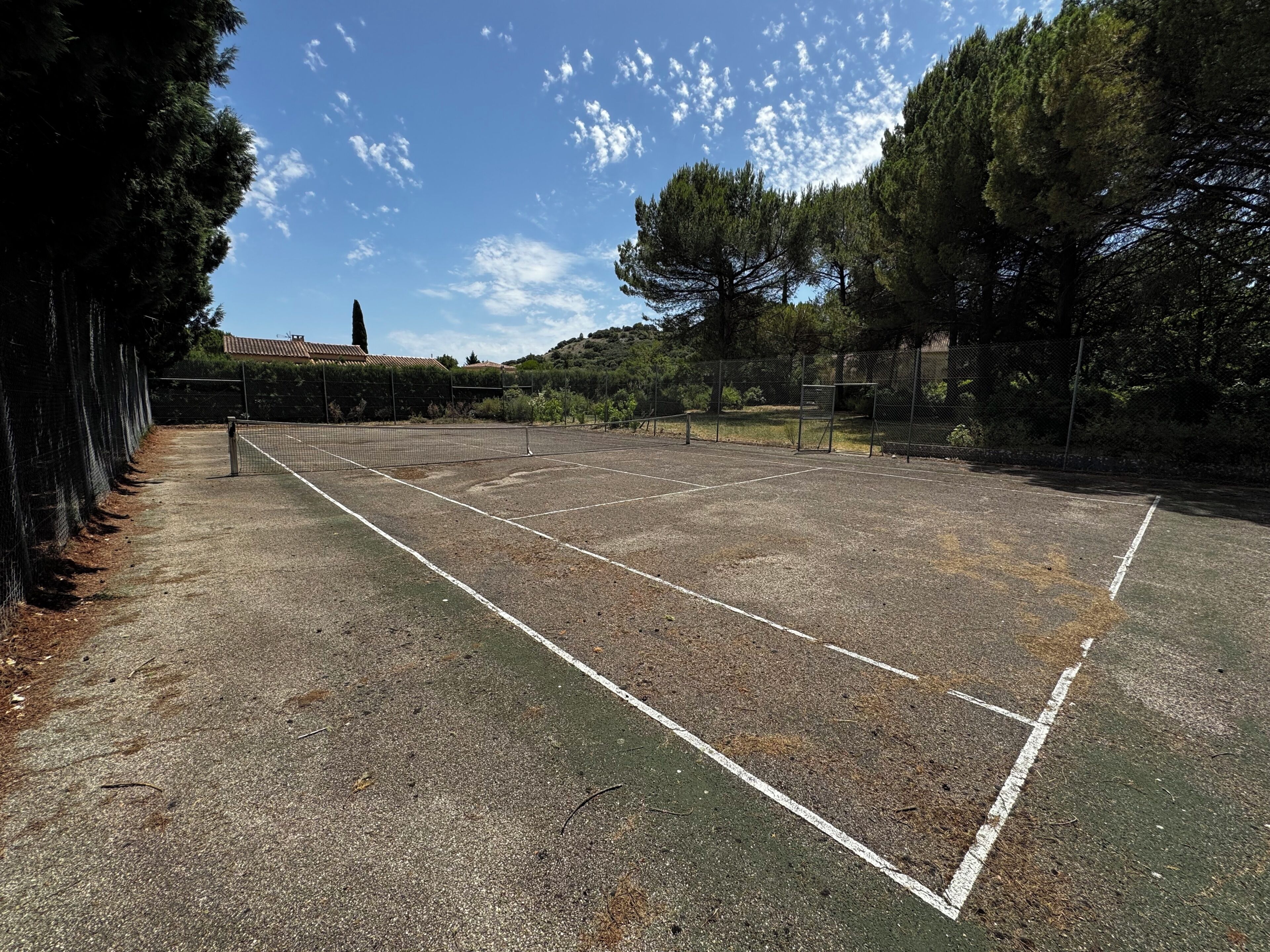 Un court de tennis en plein air, doté d’un filet, entouré d’arbres et délimité par une clôture.