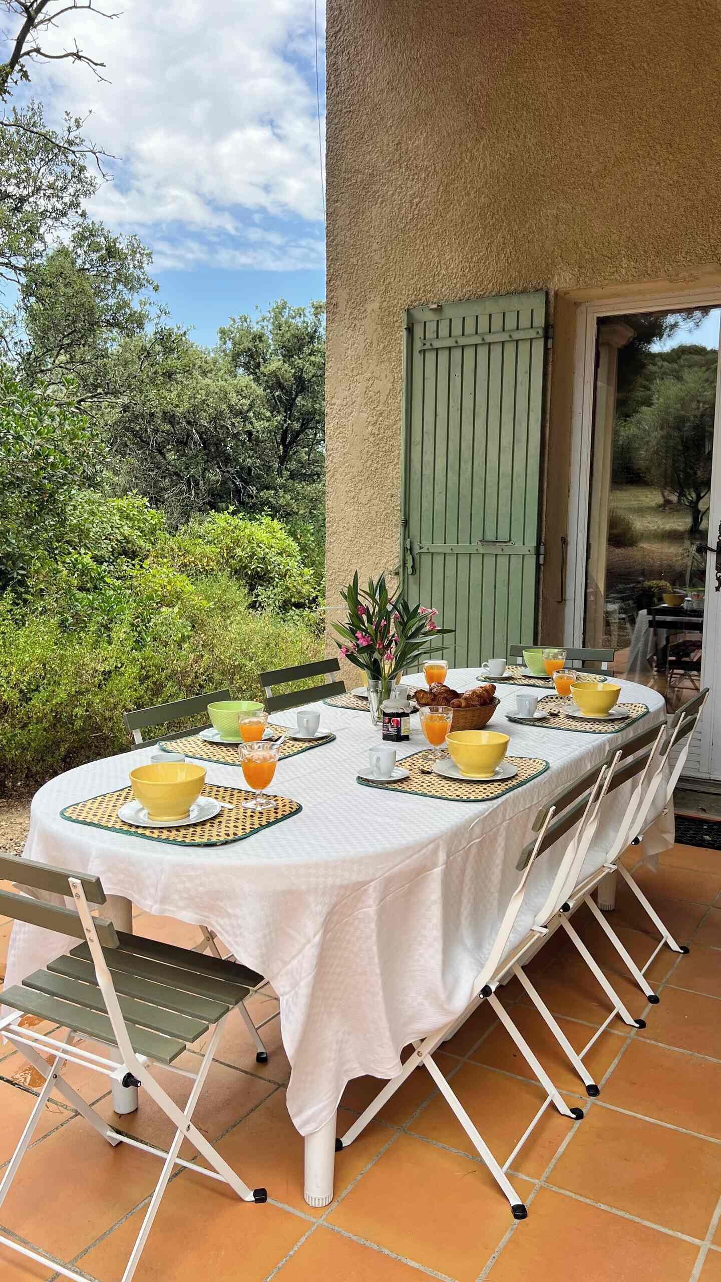 An outdoor dining area with a table set for breakfast, surrounded by greenery and a building with a green shutter.