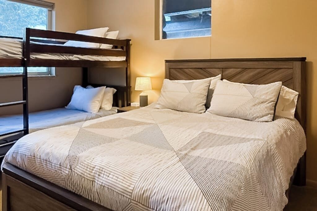 A room with a bunk bed, a single bed with a patterned bedspread, a nightstand with a lamp, and a window with a view.