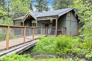 Eine Holzhütte mit einer Holzterrasse und einer kleinen Brücke, umgeben von dichtem Grün.
