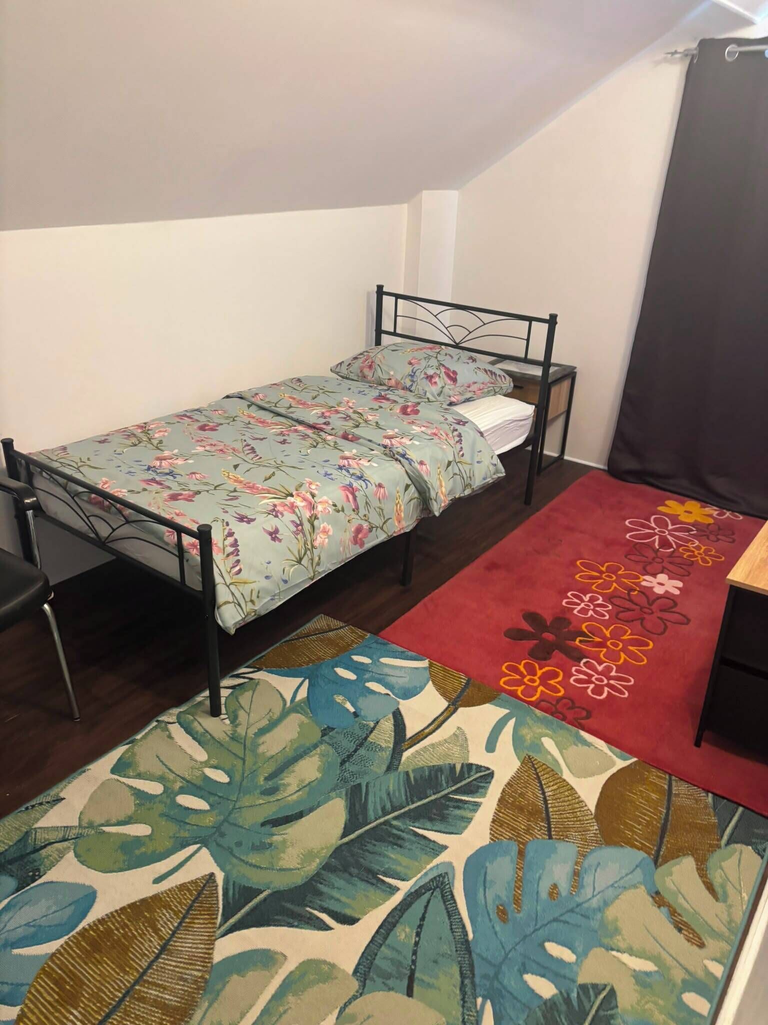 Een kleine eenpersoonskamer met een metalen bedframe, een bloemenbedekker, een rood vloerkleed met bloemenmotieven en een groot vloerkleed met groene bladeren.