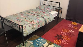Een kleine eenpersoonskamer met een metalen bedframe, een bloemenbedekker, een rood vloerkleed met bloemenmotieven en een groot vloerkleed met groene bladeren.
