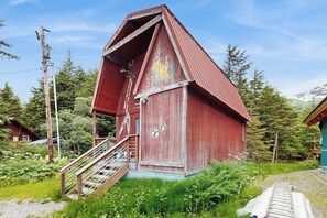 Una estructura roja de madera con una escalera que lleva a una puerta, rodeada de vegetación y un cielo despejado.