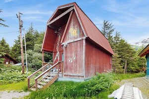 A red wooden structure with a staircase leading to a door, surrounded by greenery and a clear sky.