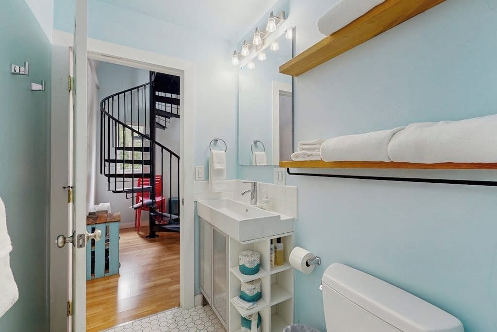 Un baño con un lavabo blanco, un espejo y un inodoro. Se ve una escalera que conduce a otra habitación.