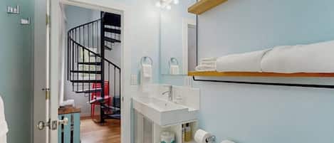 Un baño con un lavabo blanco, un espejo y un inodoro. Se ve una escalera que conduce a otra habitación.