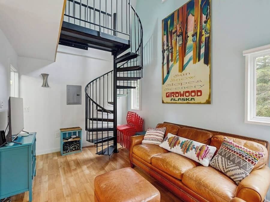 A living room with a brown leather sofa, a teal cabinet, a red chair, and a black spiral staircase.