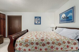 Un dormitorio con cama, mesita de noche, lámpara, puerta y un cuadro en la pared.