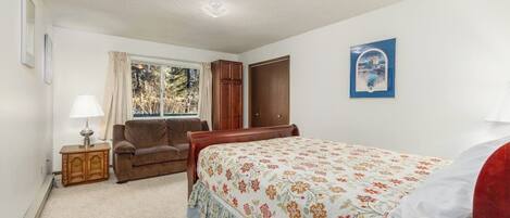 A bedroom with a bed, a wooden wardrobe, a window with curtains, a bedside table with a lamp, and a framed picture on the wall.