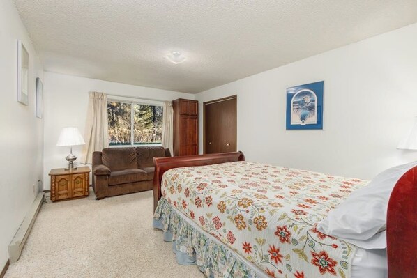 A bedroom with a bed, a wooden wardrobe, a window with curtains, a bedside table with a lamp, and a framed picture on the wall.